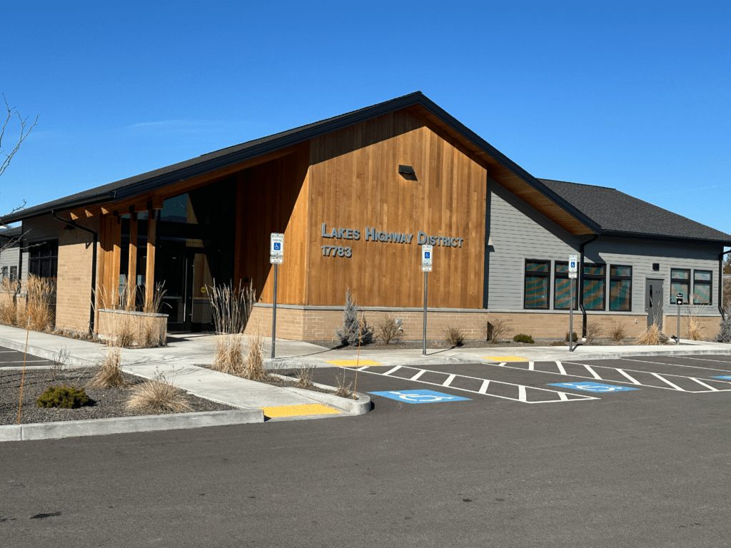 Single-story building with “Lakes Highway District 11783” on the wood facade, large windows, and accessible parking spaces in front. Landscaping includes grasses and shrubs under a clear blue sky.