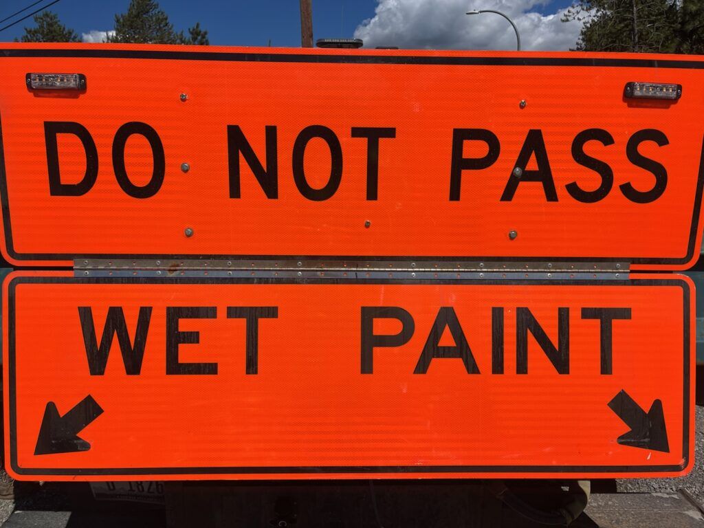 Two bright orange road signs serve as a public notice; the top sign says "DO NOT PASS" in black letters, while the bottom reads "WET PAINT" with black arrows pointing left and right, indicating ongoing road striping.